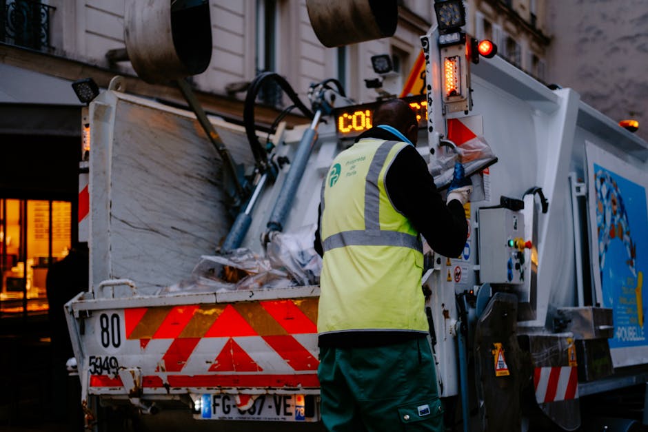 A waste collection operative wearing a high-visibility yellow vest with reflective strips and green work trousers is operating a large, white industrial refuse collection vehicle parked on a city street during evening hours. The vehicle’s rear is open, revealing a load of mixed waste, including papers and household rubbish, visible inside the open container. The worker is standing on the right side of the vehicle, using a control panel or buttons mounted on the side of the truck to manage the mechanical loading process. The vehicle features red and white chevron reflective markings on the rear for safety, and various lights, including warning and indicator lights, are illuminated. In the background, there is a building with illuminated windows, suggesting active urban surroundings, and ambient light indicates dusk or early evening, creating a slightly dim environment suited for private waste collection operations. This scene aligns with independent rubbish removal services often used in non-council waste disposal, highlighting the practical aspects of on-site waste collection undertaken by Waste Disposal Uxbridge, especially for commercial or residential clients requiring tailored rubbish clearance.