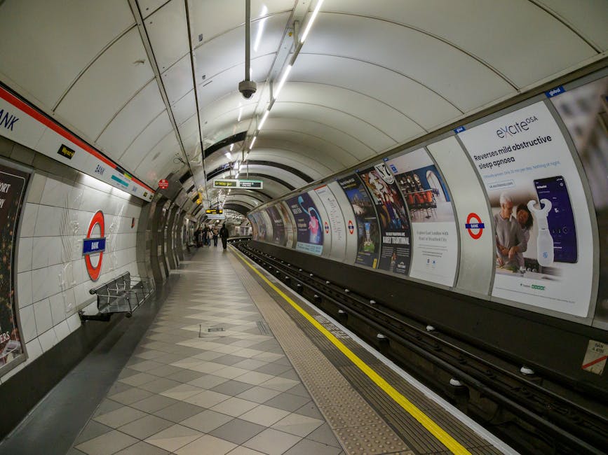 An underground London Underground station platform, featuring a curved ceiling with bright linear lighting fixtures. The platform walls are tiled with white glossy tiles, and prominent London Underground signage with a red and blue roundel is visible on the left side. A few passengers are walking along the platform, some near the far end, while others are standing closer to the wall. Along the right side, there is a series of advertising posters framed by metallic panels, displaying various products and promotional images associated with modern technology and health. The tracks run centrally, with a bright yellow safety line along the edge of the platform. The environment appears clean and well-lit, with a neutral, professional atmosphere typical of urban transit stations. This scene highlights the importance of proper waste management and disposal, as public transportation spaces often have dedicated rubbish and recycling bins to maintain cleanliness, which Waste Disposal Uxbridge offers through independent waste collection services in the area.