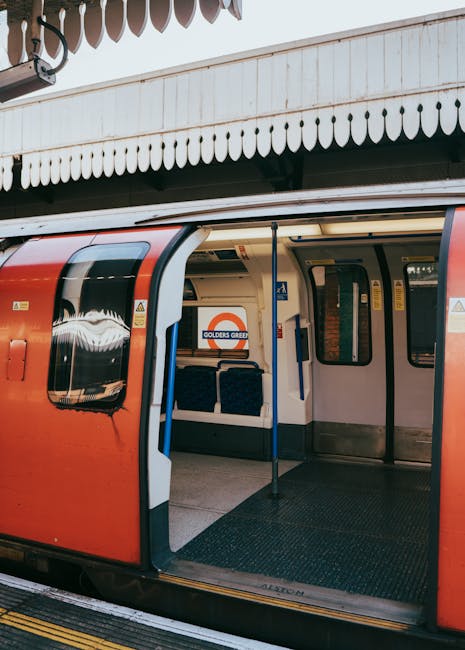Open doors of a London Underground train parked at Uxbridge station, with the station's white wooden canopy featuring decorative scalloped edging visible above. The train exterior is predominantly red with large, tinted windows reflecting light, while the interior is partially visible through the open carriage doors, revealing dark padding on the seats and yellow instructional signage inside. The platform floor is made of textured, slip-resistant material, and a blue pole supports the doorway. The background includes a partial view of the station platform and the station's signage, indicating proximity to Golden Green. The scene is evenly lit with natural light, highlighting the details of the train's smooth, shiny surface and surrounding elements, illustrating an example of independent public transport infrastructure that can relate to non-local authority arrangements for transportation and access to waste disposal services like those offered by Waste Disposal Uxbridge.