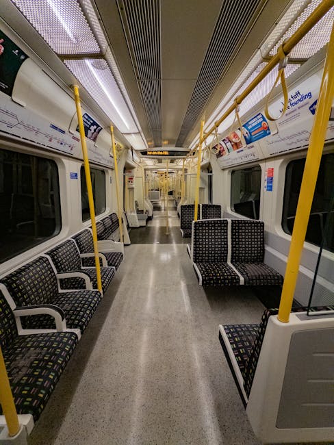 An underground London Underground station platform, featuring a curved ceiling with bright linear lighting fixtures. The platform walls are tiled with white glossy tiles, and prominent London Underground signage with a red and blue roundel is visible on the left side. A few passengers are walking along the platform, some near the far end, while others are standing closer to the wall. Along the right side, there is a series of advertising posters framed by metallic panels, displaying various products and promotional images associated with modern technology and health. The tracks run centrally, with a bright yellow safety line along the edge of the platform. The environment appears clean and well-lit, with a neutral, professional atmosphere typical of urban transit stations. This scene highlights the importance of proper waste management and disposal, as public transportation spaces often have dedicated rubbish and recycling bins to maintain cleanliness, which Waste Disposal Uxbridge offers through independent waste collection services in the area.