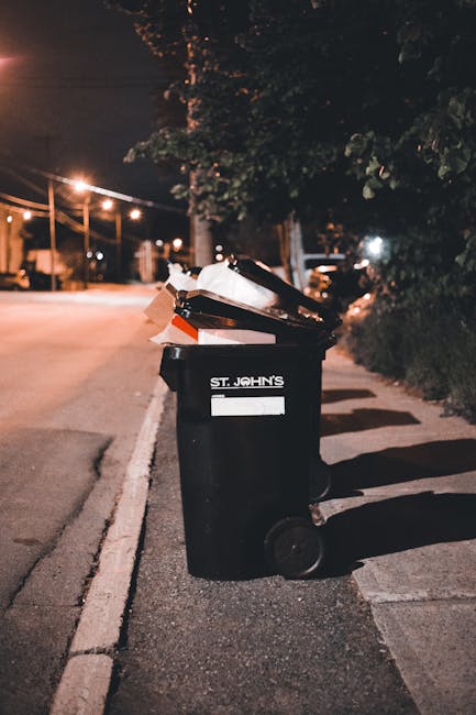 A black wheeled rubbish bin with a white label and the words 'St. John's' printed on the side, positioned on a sidewalk beside a dark road at night. The bin lid is open, revealing an overflowing collection of mixed waste, including paper, cardboard, and plastic bags. The surrounding environment features streetlights illuminating the pavement and road, with trees and shrubbery visible in the background. The scene suggests an informal or private rubbish collection context, with Waste Disposal Uxbridge providing possible alternative waste handling services for residential or commercial waste removal outside of local authority bin collections.