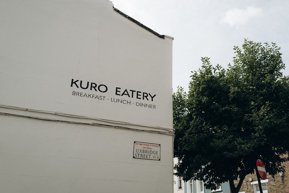 A white exterior wall of a building features a large, black, uppercase sign displaying the name 'KURO EATERY' followed by the words 'BREAKFAST - LUNCH - DINNER' underneath. To the right, there is a green leafy tree with dense foliage partially covering the sky, which appears overcast. Below the signage, mounted on the wall, is a street sign indicating 'UXBRIDGE STREET W8' with a smaller, partially visible red and white circular traffic sign beneath it. The building's simple facade and the street sign suggest a central urban area. The overall scene portrays a calm, daytime setting on a quiet street with no visible activity or crowds, emphasizing the straightforward appearance of the eatery and surrounding environment as typical of a residential or mixed-use street in Uxbridge, London. This visual context subtly aligns with the theme of local business premises possibly involved in waste management or rubbish removal services by Waste Disposal Uxbridge, who may handle waste collection for such establishments.